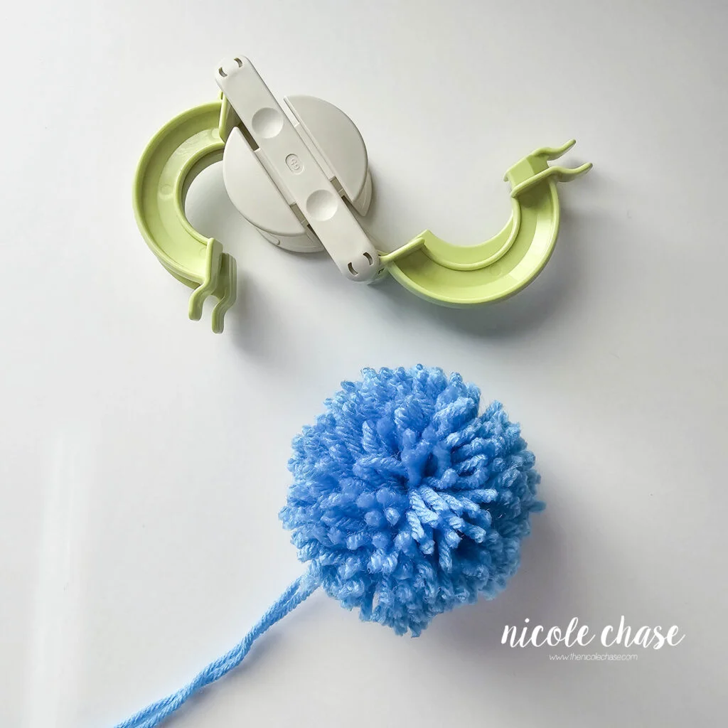 The finished blue pom pom for the free earflap hat crochet pattern next to the open pom maker. 